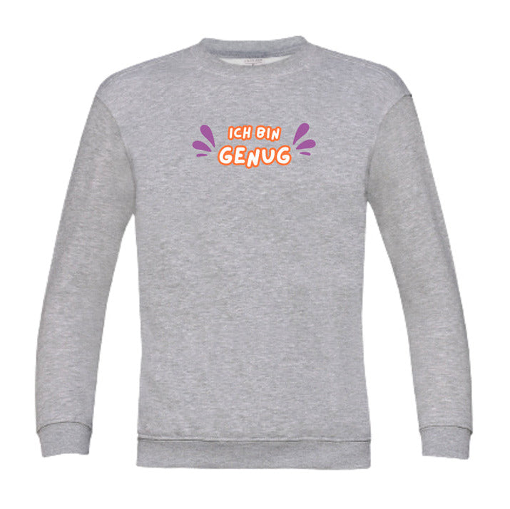 Graues Kinder-Sweatshirt mit dem Aufdruck „Ich bin genug“ in farbenfroher Schrift. Das Wort „Ich bin“ ist in orangefarbener Schrift, „Genug“ in dicker, oranger Umrandung mit weißer Füllung. Links und rechts vom Text befinden sich lilafarbene, tropfenförmige Akzente. Das Sweatshirt liegt flach auf einer weißen Oberfläche, dekoriert mit Baumwollzweigen und Eukalyptusblättern.