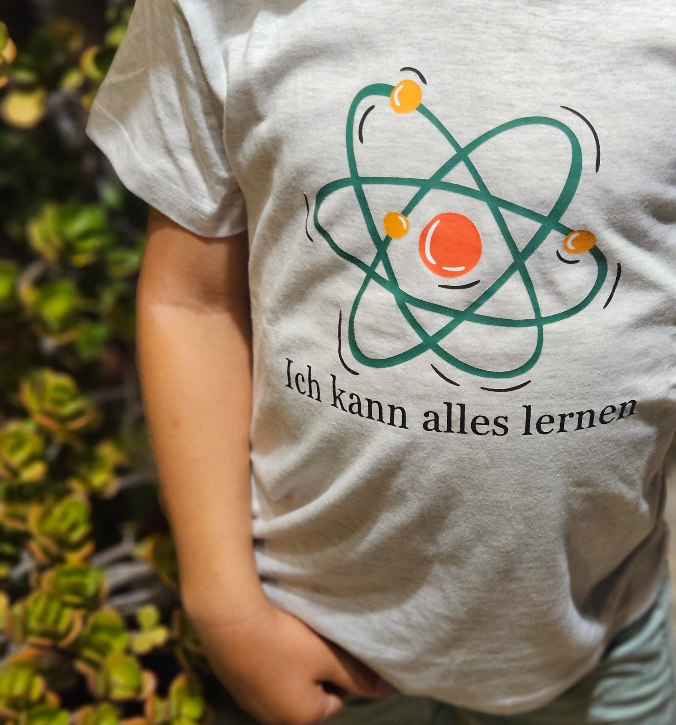  graues Kindershirt mit einem pastellfarbenen Atom-Symbol. Der Spruch „Ich kann alles lernen“ ist in schwarzer Schrift darunter gedruckt.