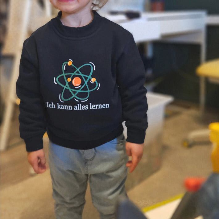navyfarbiges Kinder-Sweatshirt mit der Aufschrift „Ich kann alles lernen“ unter einer stilisierten bunten Atom-Grafik. Das Design vermittelt Neugier, Lernfreude und Wachstum.