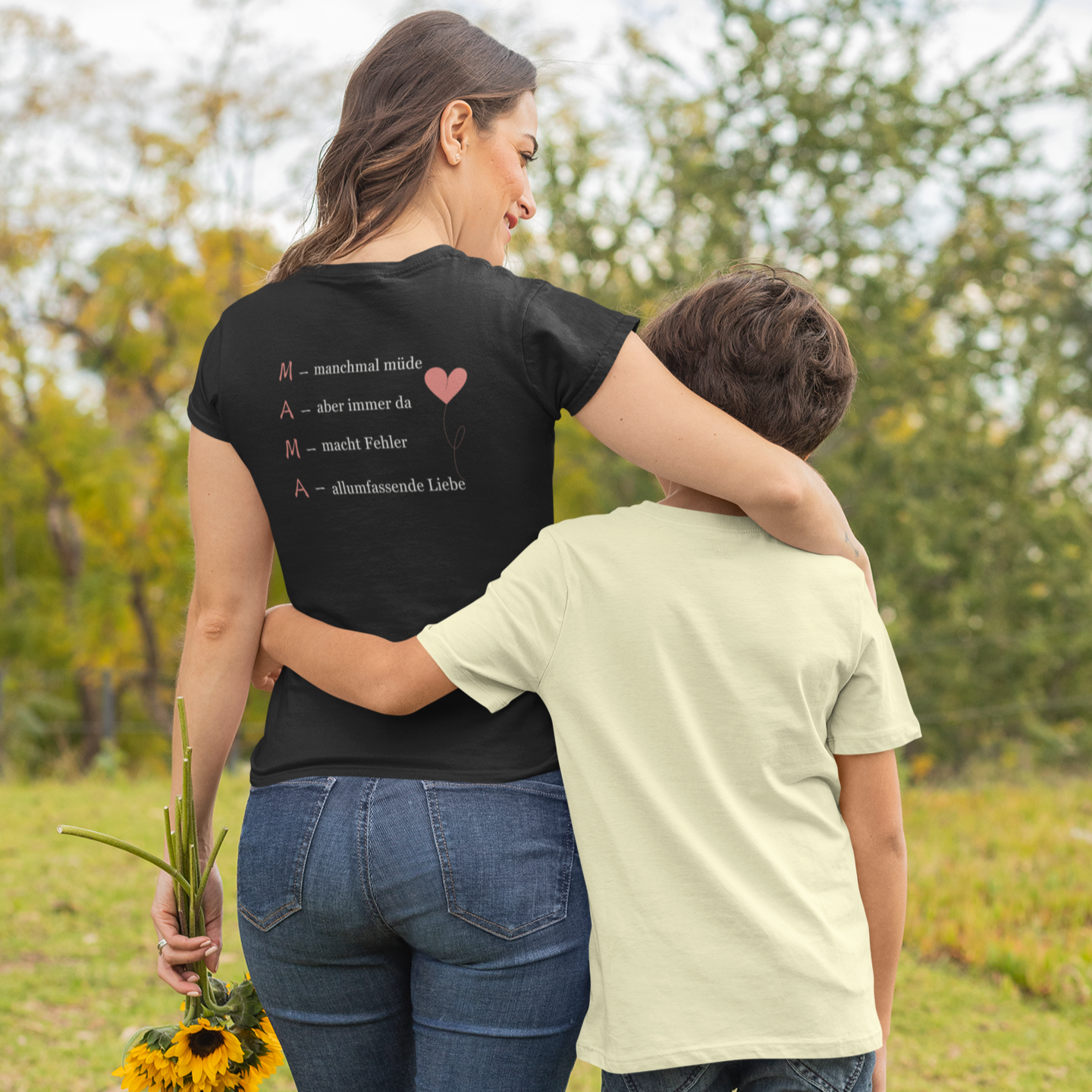 Mama mit Kind Arm in Arm auf einer Wiese, trägt schwarzes T-Shirt mit liebevollem Backprint über Mutterschaft aus der Kollektion „Achtsame Mama“ vom Sinachtsam-Shop