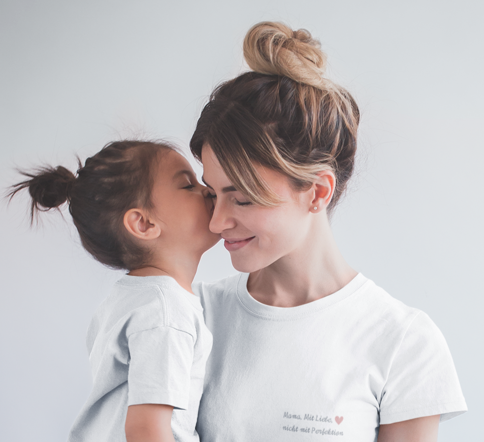 Mutter in weißem Statement-T-Shirt mit Aufdruck „Mama. Mit Liebe. Nicht mit Perfektion.“ hält ihr Kind auf dem Arm – liebevolle Szene aus der „Achtsame Mama“-Kollektion vom Sinachtsam-Shop