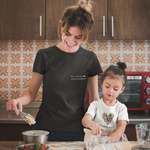 Mutter-Tochter-Moment beim Backen: Mama trägt ein schwarzes T-Shirt mit dem Aufdruck „Mama. Mit Liebe. Nicht mit Perfektion.“, Tochter trägt ein Herz-Shirt – authentische Szene aus dem Sinachtsam-Shop