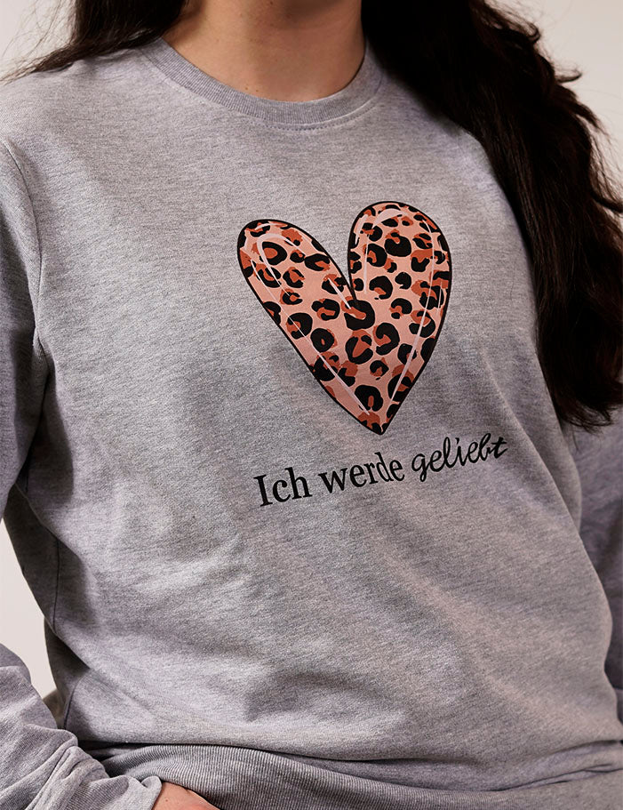 eine nahaufnahme von einer frau in einem hell grauen pullover für selbstakzeptanz mit einem großen herz und leoparden print drin mit der schwarzen schrift ich werde geliebt 