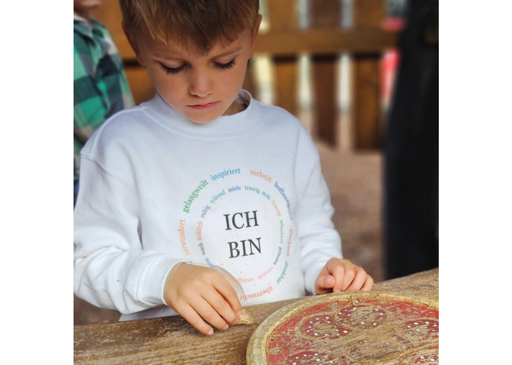 junge in einem weißen pullover mit schwarzer schrift ich bin und im kreis drumherum bunte worte wie gelangweilit, inspiriert, verletzt, hoffnungsvoll etc.