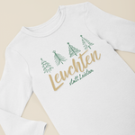 Weißer Baby-Langarmbody mit grünen Tannenbäumen und goldenem Schriftzug „Leuchten statt Leisten“, flach auf hellem Hintergrund präsentiert.