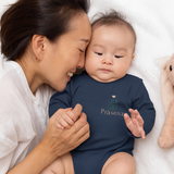 Mama kuschelt mit ihrem Baby, das einen navyfarbenen Body mit kleinem Frontprint „Präsenz“ und Geschenk-Icon trägt.