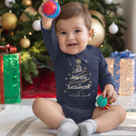 Baby in navyfarbenem Weihnachts-Body mit dem Spruch „Ich bin ein Geschenk – mit Liebe verpackt“ sitzt lachend vor einem Weihnachtsbaum und hält Spielzeug in der Hand.