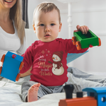 Baby trägt rotes Kinder-Longsleeve mit Schneemann-Motiv und der Affirmation „Ich bring Magie in den Winter“, weihnachtliches Motiv für kleine Kinder.