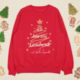Rotes Weihnachtssweatshirt mit goldener Tannenbaum-Grafik und dem Schriftzug „Ich bin ein Geschenk – mit Liebe verpackt“, umgeben von Lebkuchenfiguren und festlicher Deko.