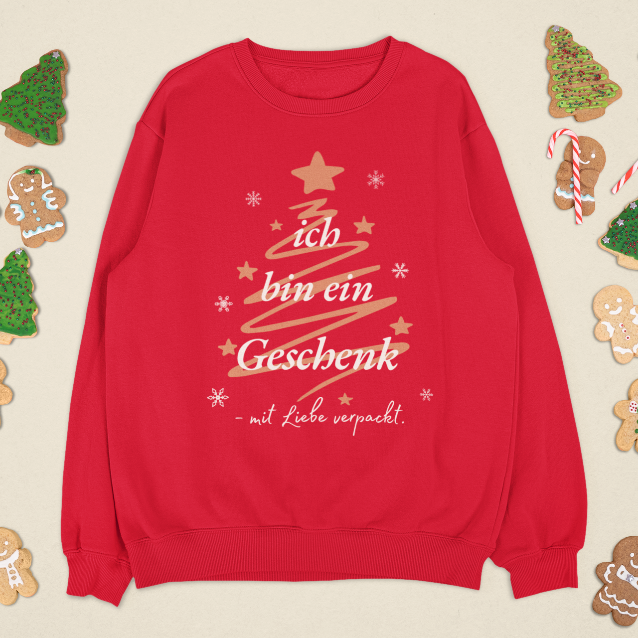 Rotes Weihnachtssweatshirt mit goldener Tannenbaum-Grafik und dem Schriftzug „Ich bin ein Geschenk – mit Liebe verpackt“, umgeben von Lebkuchenfiguren und festlicher Deko.