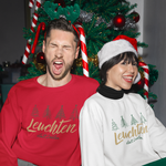 Paar in weihnachtlicher Atmosphäre vor dem geschmückten Baum, beide tragen „Leuchten statt Leisten“ Affirmations-Sweatshirts in Rot und Weiß.