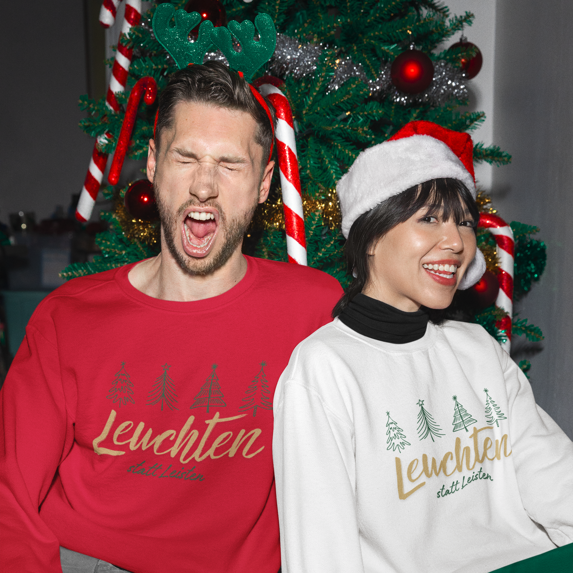 Paar in weihnachtlicher Atmosphäre vor dem geschmückten Baum, beide tragen „Leuchten statt Leisten“ Affirmations-Sweatshirts in Rot und Weiß.