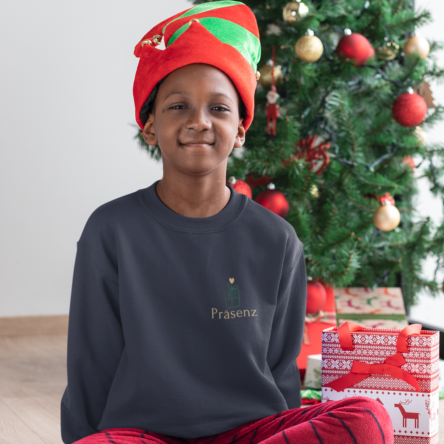 Kind sitzt vor einem Weihnachtsbaum und trägt ein dunkelblaues Sweatshirt mit kleinem Herz-Geschenk-Symbol und dem Schriftzug ‚Präsenz‘ auf der Herzseite.