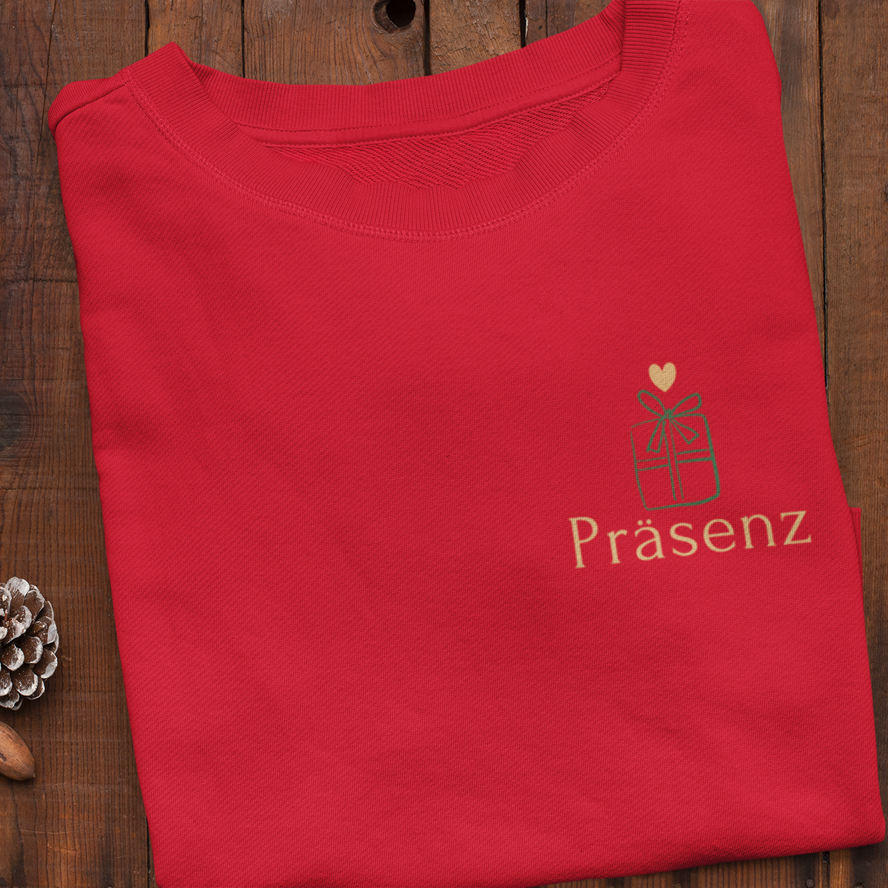 Gefaltetes rotes Sweatshirt mit kleinem Geschenk-Symbol und dem goldenen Schriftzug ‚Präsenz‘ auf der Herzseite, umgeben von weihnachtlichen Dekoelementen.