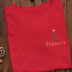 Gefaltetes rotes Sweatshirt mit kleinem Geschenk-Symbol und dem goldenen Schriftzug ‚Präsenz‘ auf der Herzseite, umgeben von weihnachtlichen Dekoelementen.